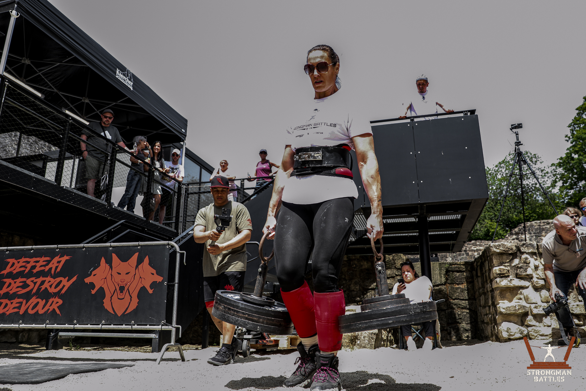 5P0A4015 Woman competing in the farmers walk with spectators and an outdoor event stage in the background